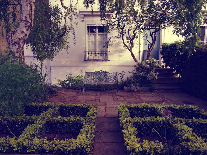 A quaint garden area featuring a paved stone patio with neatly trimmed low hedges forming flower bed borders in the foreground. Central to the patio is an ornate wrought-iron bench placed against a wh